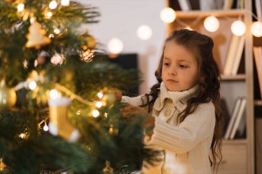 Örgü kazaklı küçük sevimli kız Noel ağacına ışık tutarak süsleme yapıyor. Güzel oturma odasında Noel ağacı süsleyen çocuk. Mutlu aile tatilleri Yeni Yıl konsepti.