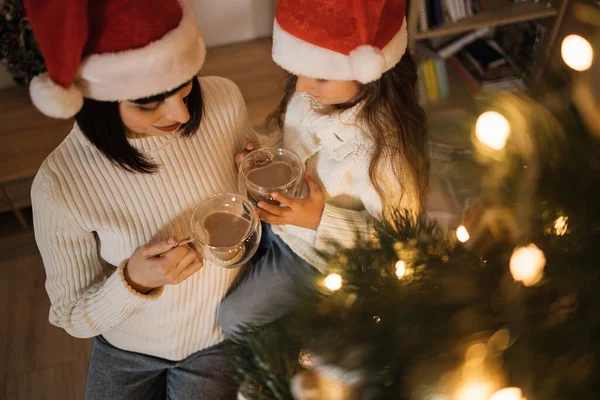 Cropped top view portrait of beautiful young caucasian mother and her cute little daughter in santa hat drinking hot cocoa or chocolate from cups near decorated Chritmas tree.