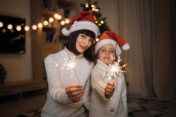 Focus on sparklers, blurred happy mother and little cute daughter holding bengal lights in their hands on background of decorated christmas tree at home.