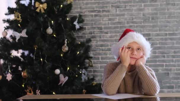 サンタに手紙を書く赤いクリスマスの帽子の少年 テーブルでクリスマスツリーの近くに座って夢と笑顔 家にいてお祝いのコンセプト — ストック動画