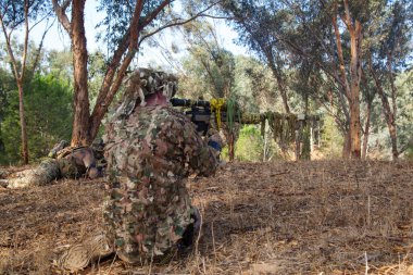 Orman bölgesinde kamuflajlı askerler. Bir asker yerde yatıyor, ikincisi dizlerinin üstüne çöküp otomatı hedef alıyor.