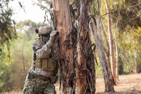 一个披着迷彩服的男人躲在一棵树后 透过双筒望远镜观看 军队对敌人进行侦察和跟踪 — 图库照片