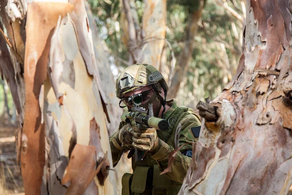 A guy in camouflage between a tree is aiming at the calimator of the machine gun. Dressed in military ammunition with a machine gun in his hands. Airsoft needle hides behind a tree and fires