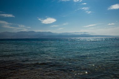 Coastal area of the Red Sea in Eilat with mountain views. Stones are visible in the water near the beach with a view of the mountains in a blue haze. Beautiful clouds in the sky in blue tones. Shine of water in the sun