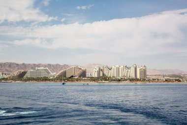 Kızıl Deniz 'deki Eilat' taki otel kompleksinin manzarası. Deniz kenarındaki güzel dağların arkasında büyük oteller. Otel kompleksinin üzerinde güneşli bir hava var. İsrail 'de bulutlu mavi gökyüzü. Otellerde dinlenin.