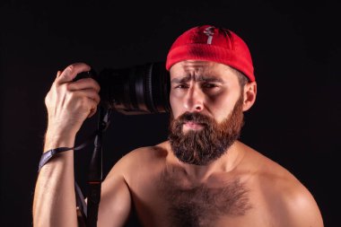 A bearded photographer on a black background aims at his temple with a camera. Tired eye of a creative person. Work burnout