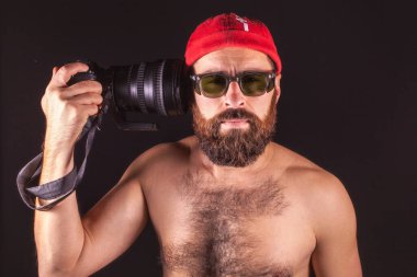 Creative photo of a photographer. The guy aims at his temple with a lens. Model in a red cap on a black background
