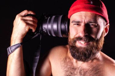 A model on a black background with a camera in her hands looks into the frame with a smile. The camera in his hand is aimed at the guy's temple, and he decreases with clenched teeth.