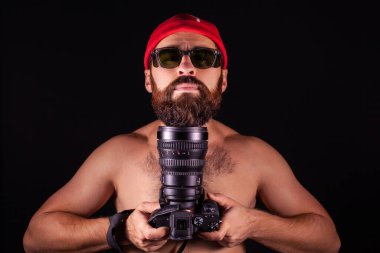 A man with a beard on a black background with a camera. The guy in the red cap puts his head on the lens.