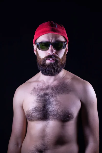 A guy with a naked torso and in a red cap stands on a black background. On his head is a red cap and green glasses. Regular looking guy with hairy chest