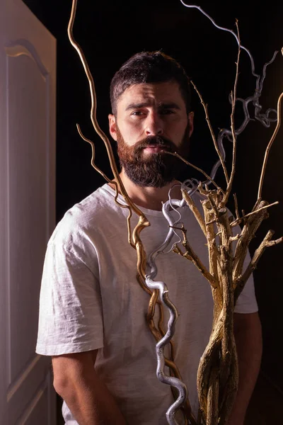 A guy with a beard in a white T-shirt is standing at the door. Decorative branches painted in gold and silver in front of the model. Handsome man