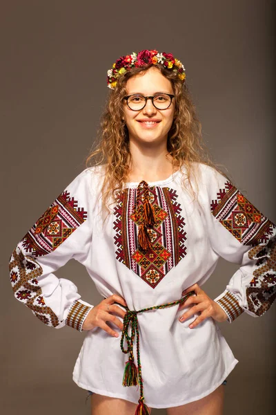 Vertical photo of a woman in an embroidered dress. The model is standing in a pose - hands out to the sides with a wreath on her head and wearing glasses. The smile of a girl in national Ukrainian clothes