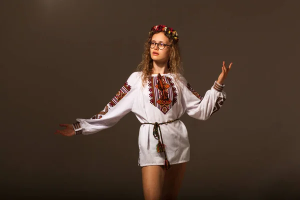 A model on a gray background in a national dress and a wreath of flowers. One hand of the girl is raised to the mountain and the other is taken to the side. A beautiful, young, blond girl in an embroidered dress