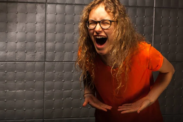 Girl screams loudly against the background of a gray wall. The emotion of anger, rage and powerlessness. A wild woman with curly hair and glasses screams. Uncontrollable pain and burnout at work.