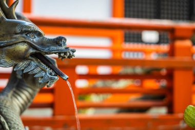 Bir su çeşmesinin yakınında ejderha kafası şeklinde bir fıskiyenin arkasında odak dışı turuncu yatay parmaklıklar var.