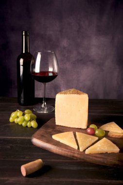 A piece of Manchego Cheese from Spain with some green and red grapes and a glass and bottle of Spanish red wine at the background in a dark food photography style