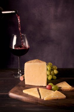 A piece of Manchego Cheese from Spain with some green and red grapes and a glass and bottle of Spanish red wine at the background in a dark food photography style