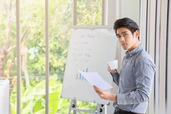 Business and Comminication Concept. Asian young businessman standing looking to camera with holding a white cup of hot coffee and paper in meeting room with whiteboard