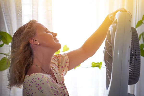 Woman cooling herself with fan. The girl escapes from the heat with a home fan at home. Exhausting summer day high temperature weathe heat. Woman enjoying air flow from fan in living room. Summer heat