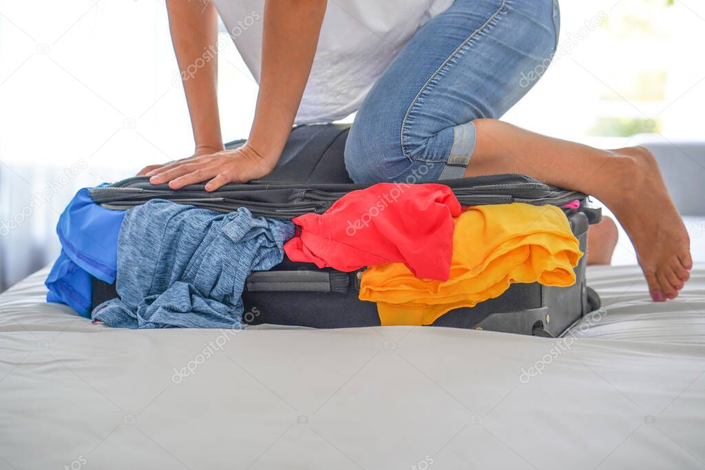 Woman closes the suitcase with her feet on bed. Young woman packing suitcase on bed. Young girl presses her foot on a crowded suitcase to close it, getting ready for the trip, vacation, travel