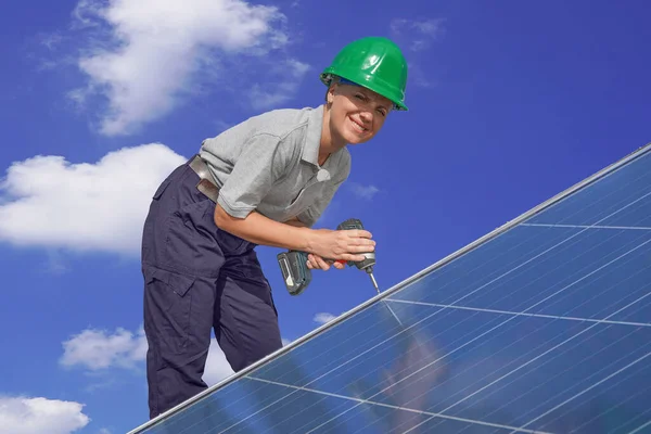 Woman engineer, electrician in green protective helmet against background of cloudy sky installing solar photovoltaic panel system using screwdriver. Professional electrician mounting solar panel