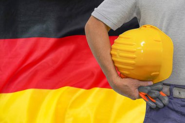 Close-up of hard hat holding by construction worker on Germany flag background. Hand of worker with yellow hard hat and gloves. Concept of Industry, construction and industrial workers in Germany
