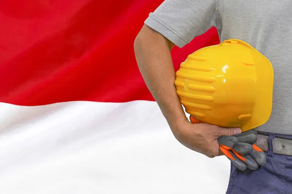 Close-up of hard hat holding by construction worker on Indonesia flag background. Hand of worker with yellow hard hat and gloves. Concept of Industry, construction and industrial workers in Indonesia