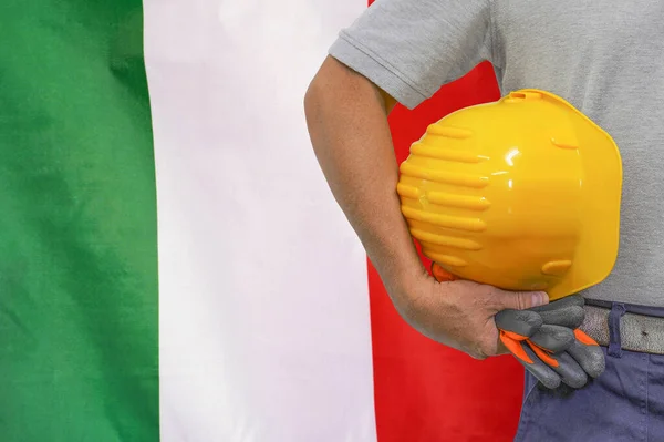 Close-up of hard hat holding by construction worker on Italy flag background. Hand of worker with yellow hard hat and gloves. Concept of Industry, construction and industrial workers in Italy