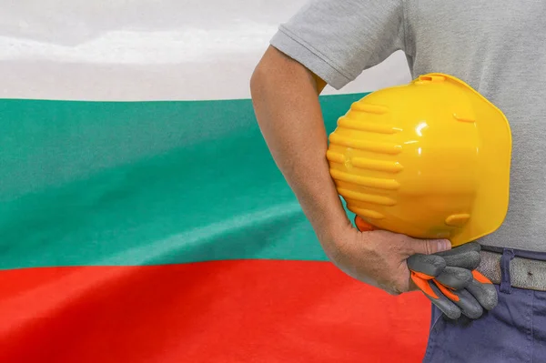 Close-up of hard hat holding by construction worker on Bulgaria flag background. Hand of worker with yellow hard hat and gloves. Concept of Industry, construction and industrial workers in Bulgaria