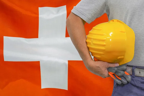 Close-up of hard hat holding by construction worker on Switzerland flag background. Hand of worker with yellow hard hat and gloves. Concept of Industry, construction and industrial workers in Swiss