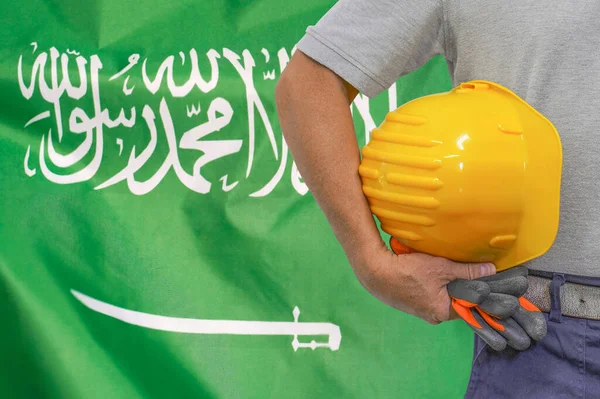 Close-up of hard hat holding by construction worker on Saudi arabia flag background. Hand of worker with yellow hard hat and gloves. Concept of Industry, construction and workers in Saudi arabia