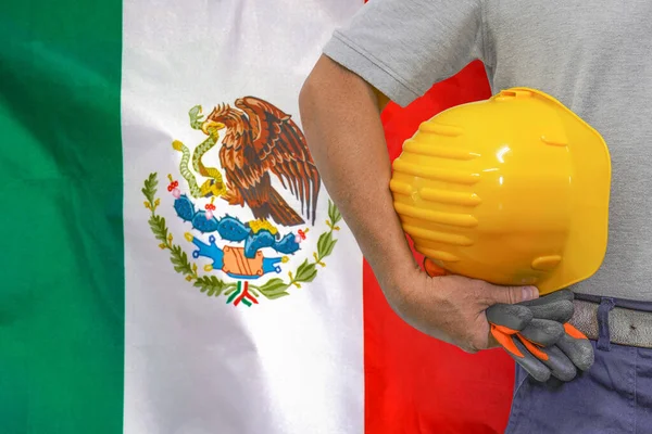 Close-up of hard hat holding by construction worker on Mexico flag background. Hand of worker with yellow hard hat and gloves. Concept of Industry, construction and industrial workers in Mexico