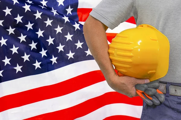 Close-up of hard hat holding by construction worker on US flag background. Hand of worker with yellow hard hat and gloves. Concept of Industry, construction and industrial workers in USA