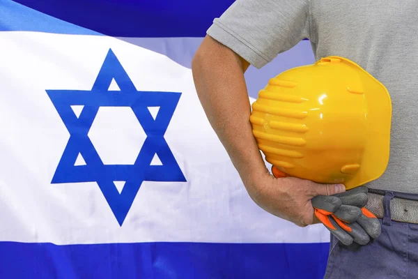 Close-up of hard hat holding by construction worker on Israel flag background. Hand of worker with yellow hard hat and gloves. Concept of Industry, construction and industrial workers in Israel