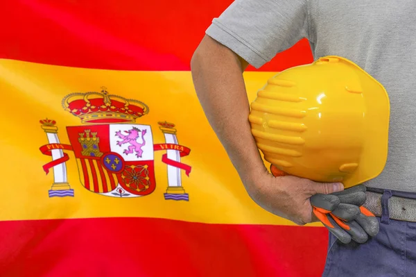 Close-up of hard hat holding by construction worker on Spain flag background. Hand of worker with yellow hard hat and gloves. Concept of Industry, construction and industrial workers in Spain