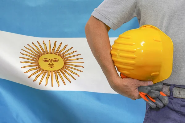 Close-up of hard hat holding by construction worker on Argentina flag background. Hand of worker with yellow hard hat and gloves. Concept of Industry, construction and industrial workers in Argentina