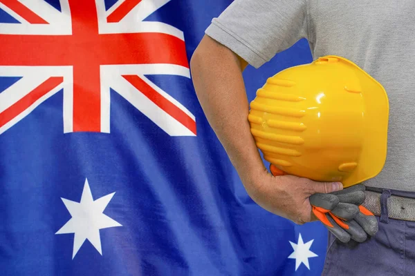 Close-up of hard hat holding by construction worker on Australia flag background. Hand of worker with yellow hard hat and gloves. Concept of Industry, construction and industrial workers in Australia
