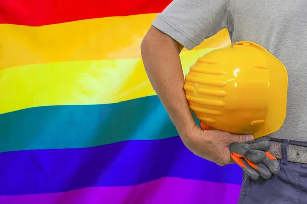 Close-up of hard hat holding by construction worker on LGBT flag background. Hand of worker with yellow hard hat and gloves. Concept of Industry, construction and industrial workers LGBT