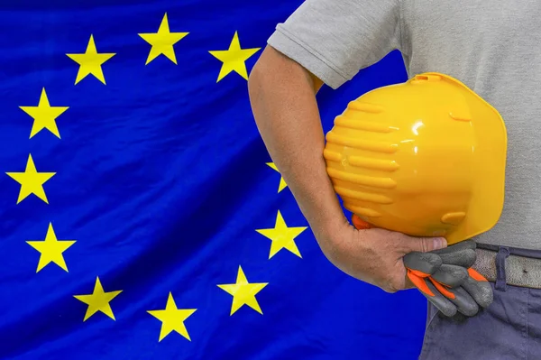 Close-up of hard hat holding by construction worker on EU flag background. Hand of worker with yellow hard hat and gloves. Concept of Industry, construction and industrial workers in European Union