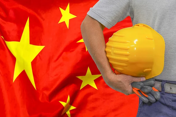Close-up of hard hat holding by construction worker on China flag background. Hand of worker with yellow hard hat and gloves. Concept of Industry, construction and industrial workers in China