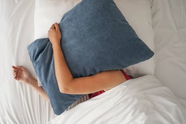Woman in bed covering herself with a pillow her head. Woman is woken up in the morning and annoyed by noise from neighbors or from the street. Stress, family quarrels. Avoiding responsibility