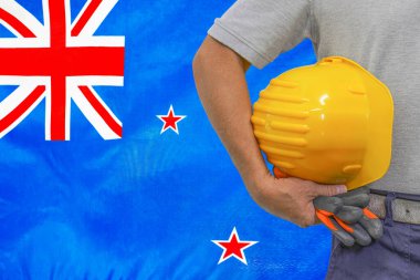 Close-up of hard hat holding by construction worker on New Zealand flag background. Hand of worker with yellow hard hat and gloves. Concept of construction and industrial workers in New Zealand