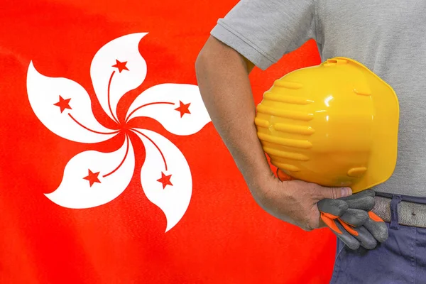 Close-up of hard hat holding by construction worker on Hong Kong flag background. Hand of worker with yellow hard hat and gloves. Concept of Industry, construction and industrial workers in Hong Kong