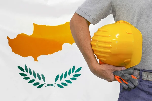 Close-up of hard hat holding by construction worker on Cyprus flag background. Hand of worker with yellow hard hat and gloves. Concept of Industry, construction and industrial workers in Cyprus