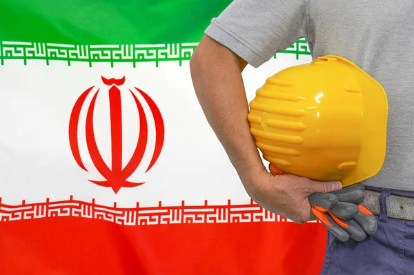 Close-up of hard hat holding by construction worker on Iran flag background. Hand of worker with yellow hard hat and gloves. Concept of Industry, construction and industrial workers in Iran