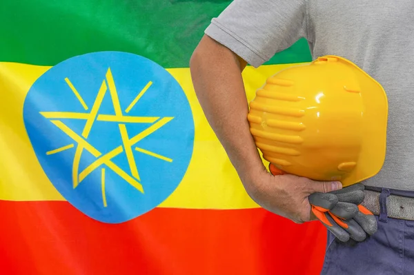 Close-up of hard hat holding by construction worker on Ethiopia flag background. Hand of worker with yellow hard hat and gloves. Concept of Industry, construction and industrial workers in Ethiopia