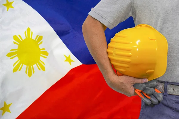 Close-up of hard hat holding by construction worker on Philippines flag background. Hand of worker with yellow hard hat and gloves. Concept of construction and industrial workers in Philippines