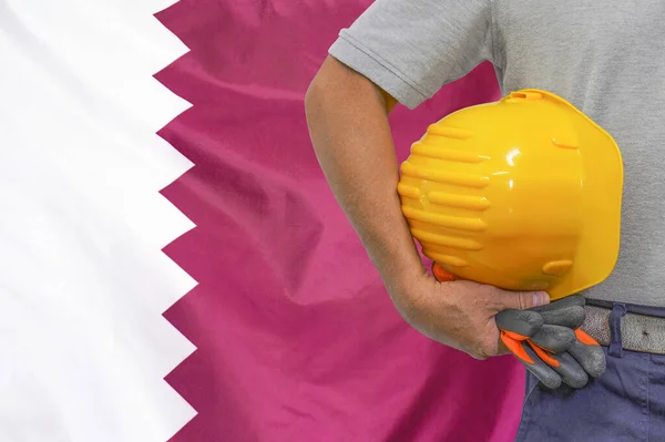 Close-up of hard hat holding by construction worker on Qatar flag background. Hand of worker with yellow hard hat and gloves. Concept of Industry, construction and industrial workers in Qatar