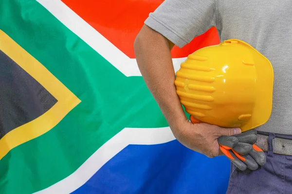 Close-up of hard hat holding by construction worker on South Africa flag background. Hand of worker with yellow hard hat and gloves. Concept of construction and industrial workers in South Africa