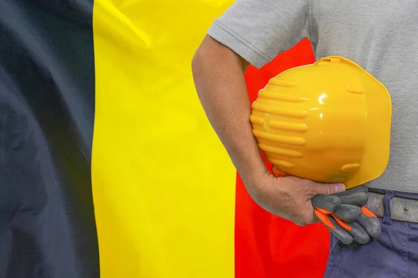 Close-up of hard hat holding by construction worker on Belgium flag background. Hand of worker with yellow hard hat and gloves. Concept of Industry, construction and industrial workers in Belgium